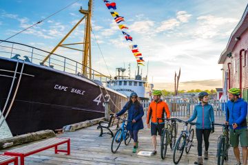 4 cyclists walking with their bikes along the boardwalk next to a restored fishing trawler berthed at Lunenburg, Nova Scotia.