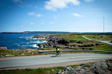 This image shows a person cycling along a scenic coastal road in Atlantic Canada, near the coastline of in the Bonavista region of Newfoundland