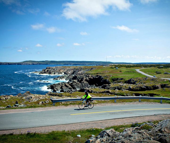 This image shows a person cycling along a scenic coastal road in Atlantic Canada, near the coastline of in the Bonavista region of Newfoundland