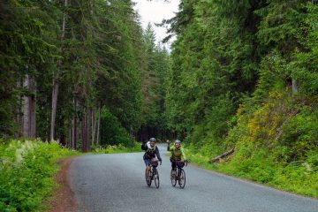 This image shows two individuals enjoying a scenic bicycle tour on a paved road surrounded by dense trees on Vancouver Island