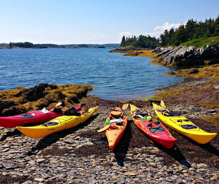 This image shows several colorful kayaks resting on a rocky shoreline in Nova Scotia, a popular activity in the area