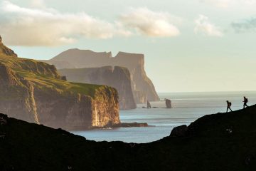The image shows two people hiking on a mountainside overlooking dramatic cliffs and the ocean of the coast of Faroe Islands