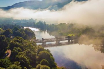 The image features a tranquil river scene in the Scottish Highlands, likely near Fort William or Ben Nevis, partially obscured by thick morning mist
