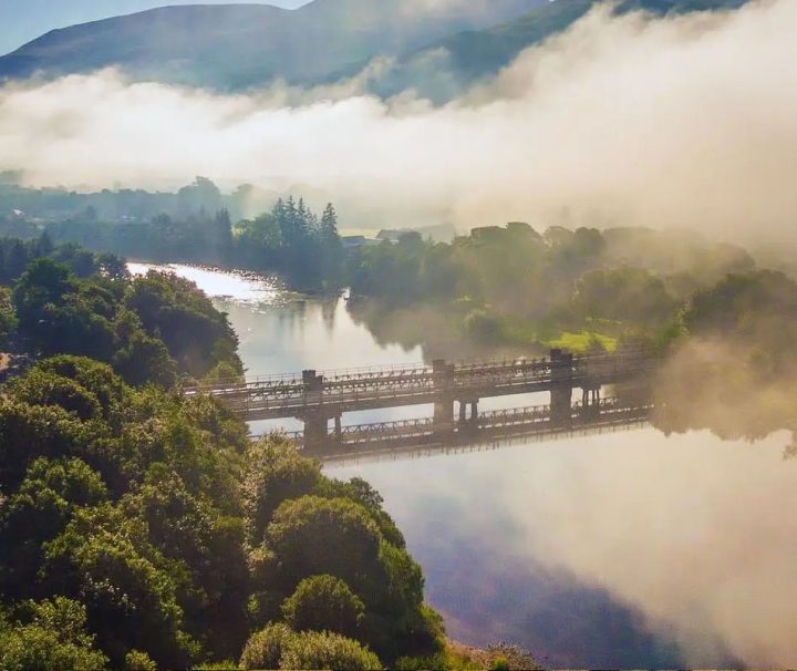 The image features a tranquil river scene in the Scottish Highlands, likely near Fort William or Ben Nevis, partially obscured by thick morning mist